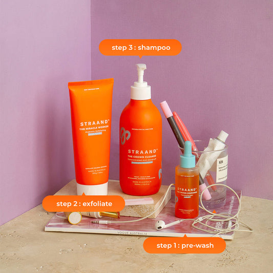 A set of three orange-colored hair care products from STRAAND, including a pre-wash scalp oil, a crown cleanse shampoo, and a miracle worker exfoliating scalp scrub.