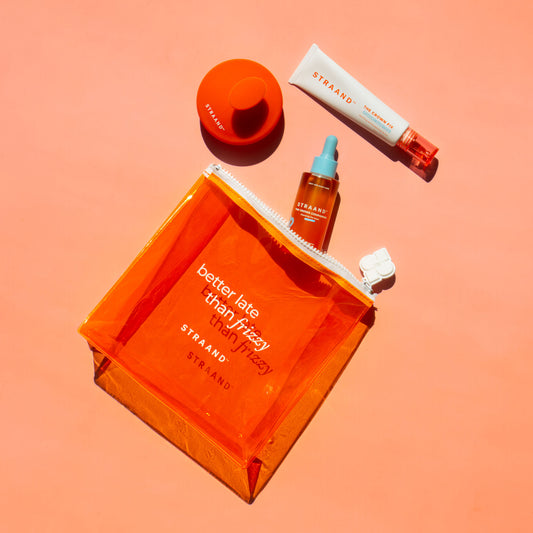 A collection of hair care products including a pre-wash scalp oil, a scalp serum, and a scalp brush, packaged in a bright orange translucent bag with 'better late than frizzy' text, and a small orange sun-shaped accessory.