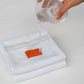 White towel with an orange label being soaked by a hand pouring water from a clear glass container.
