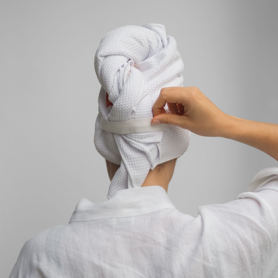 Person with a towel wrapped around their head applying cream to their face against a gray background